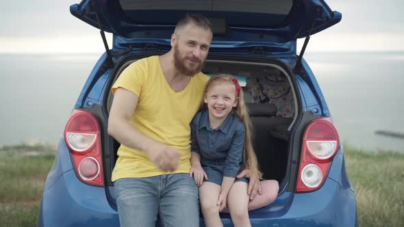 Cheerful Caucasian Bearded Father and Cute Little Daughter Sitting in Car Trunk Talking Looking Away