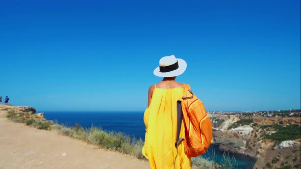 Camera Following Girl Traveler in Orange Sundress and Hat Walking To Precipitous Cliff Edge