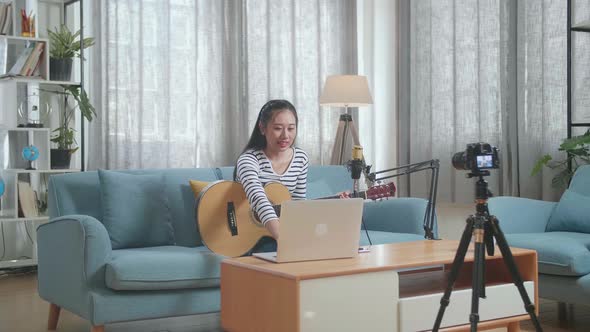 Asian Woman Reading Comments On Laptop And Speaking To Camera While Live Stream Playing A Guitar