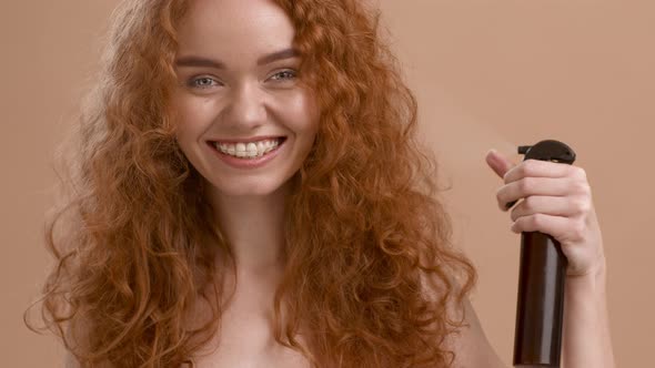 RedHaired Woman Spraying Hairspray Styling Curly Hair On Beige Background