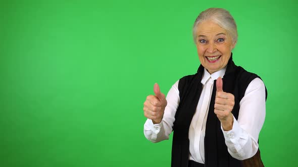 An elderly woman smiles and shows a double thumb up to the camera - green screen studio