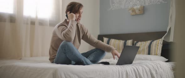 A man talking to someone on the phone while working with a laptop from home.