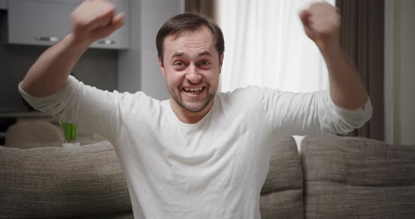 Man Rejoices Shouting with Happiness Victory Raises Hands Looks at Camera Sitting on Sofa at Home in