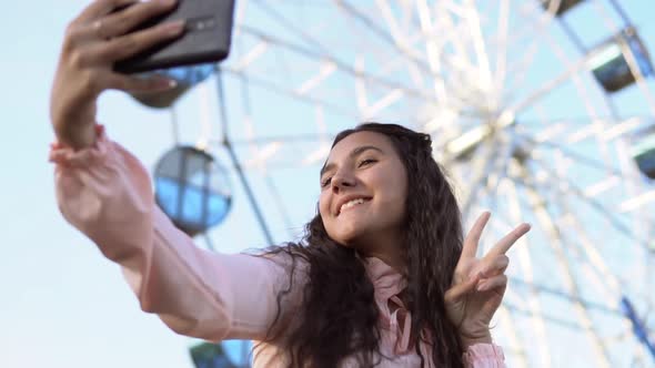 a Girl with Long Hair in a Dress Makes Selfie Using a Phone Standing Near the Ferris Wheel