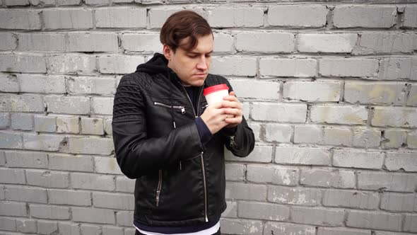 Young guy drinking coffee, against a brick wall, wearing a black leather jacket