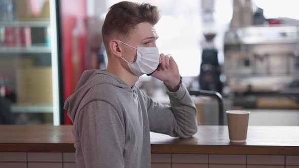 Side View of Young Man in Coronavirus Face Mask Talking on the Phone Standing at Cafe Counter with