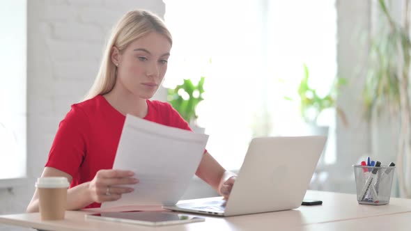 Young Blonde Woman Reading Documents and Using Laptop at Work