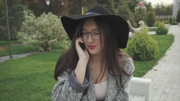 Young Woman in Black Hat and Glasses Talking on the Mobile Phone in a City Park