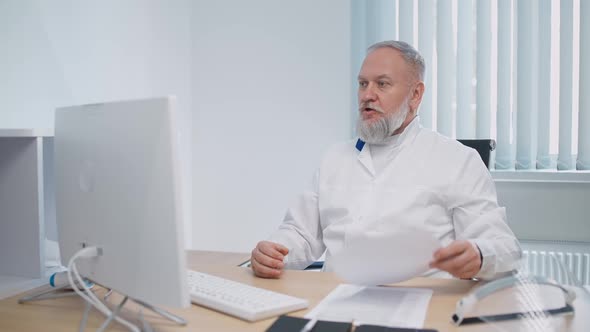 Grayhaired Male Doctor Sits at Workplace and Communicates with Colleagues Via Video Call Remote