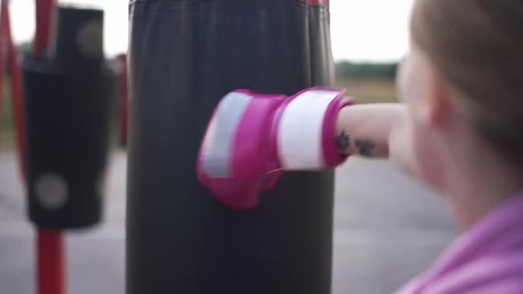 Punching Bag Hanging Outdoors with Tattooed Female Hand in Boxing Gloves Kicking Equipment in Slow