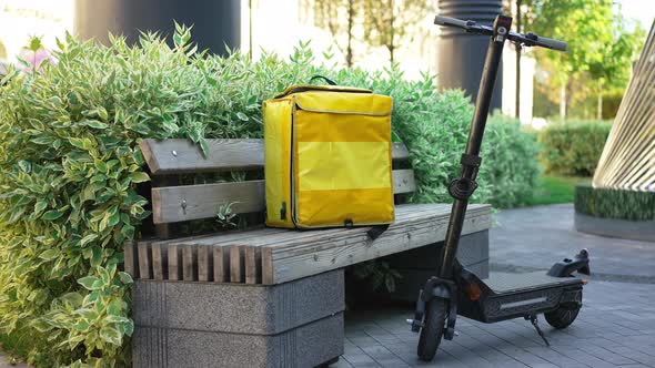 Wide Shot Electric Scooter and Yellow Food Delivery Bag on Bench on Urban City Street