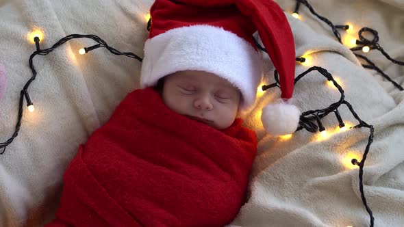 Top View Portrait First Days Of Life Newborn Cute Funny Sleeping Baby In Santa Hat Wrapped In Red