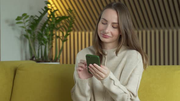 Woman Uses a Smartphone While Sitting on a Yellow Sofa in Her Living Room