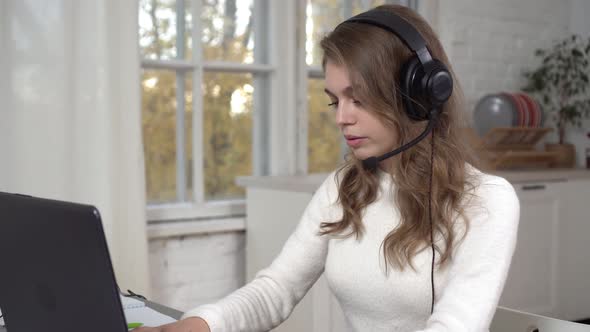 A Young Woman in Headphones with a Headset Works at a Laptop