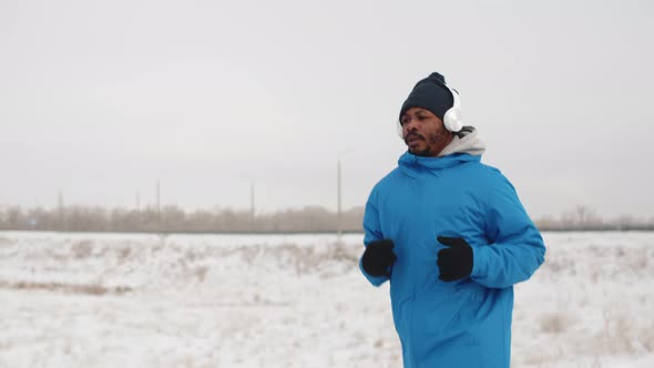 Black Man in Headphones and Warm Winter Clothes is Jogging Outdoors Front View