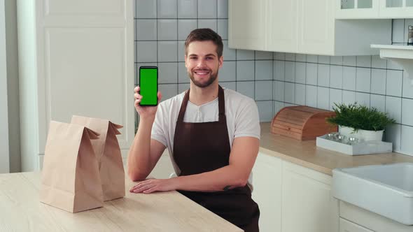 A Man Holds a Smartphone with a Green Screen in His Hand and Smiles While Sitting in the Kitchen