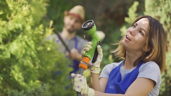 Portrait of Attractive Caucasian Woman Playing with Water Hose in the Foreground While Man Cutting