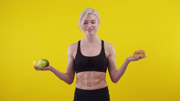 Sports woman thinks and holds fruit and burger in hands, choosing fast food, in studio.