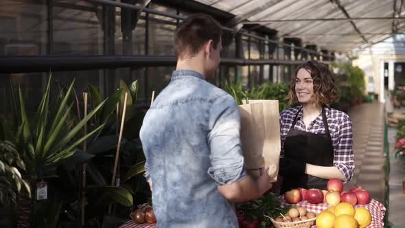 Cheerful Saleswoman in Black Apron Giving Fresh Vegetables in Paper Bag to Customer in Greenhouse