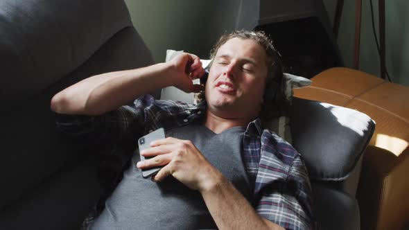 Relaxed caucasian man using smartphone and wearing headphones lying on sofa in living room