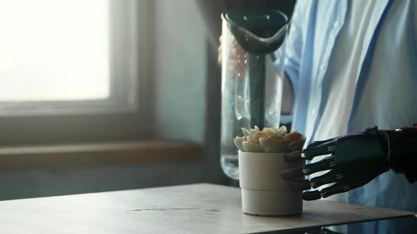 Disabled guy cyborg with bio hand prothesis pours water into green pot plant on kitchen table