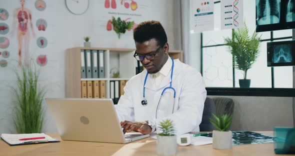 African Male Medical Worker in Eyeglasses Working on Laptop in Modern Clinic workroom
