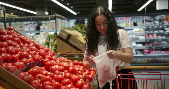 Young Woman in the Grocery Section of a Supermarket Picks Up a Bag and Puts Tomatoes in a Basket to