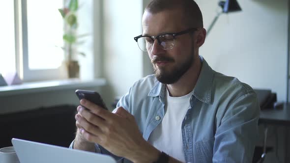 Handsome Guy is Using Smartphone at Table with Laptop in Modern Interior