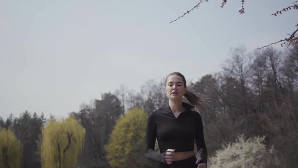 Slim Young Woman in Sportswear Running on the Riverbank