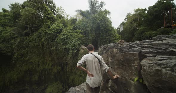 Close Up Shot of a Young Male Traveler Standing on the Stone Cliff Edge Overlooking a Waterfall in