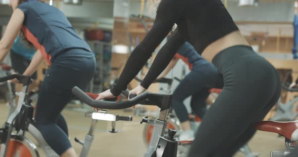 Side View of Cheerful Young Women Training on Exercise Bikes with Help of Coach. Slim Athletic alt