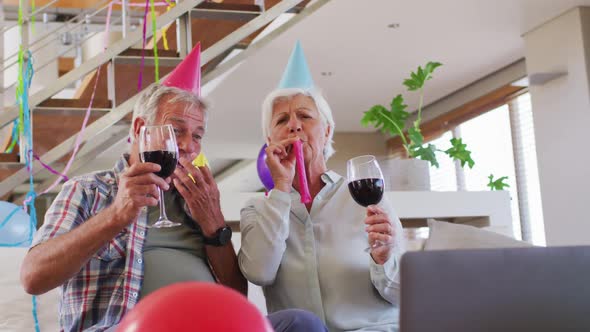 Senior caucasian couple blowing party blowers celebrating birthday toasting wine glasses at home