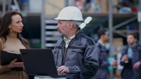 Concentrated Busy Senior Warehouse Manager in Hard Hat Checking Orders on Laptop As Young Beautiful