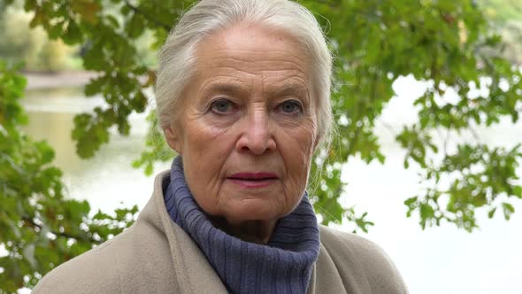 An Elderly Woman Looks Seriously at the Camera - Face Closeup - a Lake in the Blurry Background