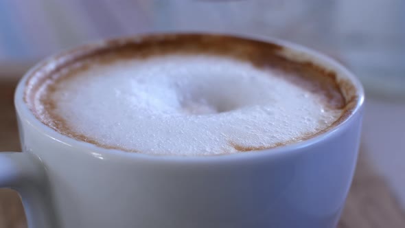 Woman stirs with a spoon the thick foam of hot cappuccino coffee in a glass cup.