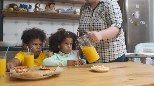 Caucasian Mother Pouring Juice in Glass for Mixed Race Children Sitting in Kitchen and Eating Pizza