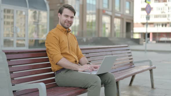 Young Man with Laptop Smiling at Camera while Sitting Outdoor on Bench