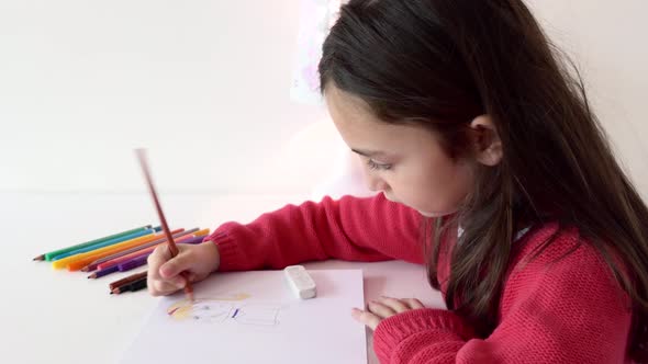 A young girl sitting at a brightly-lit table coloring with a brown colored pencil.