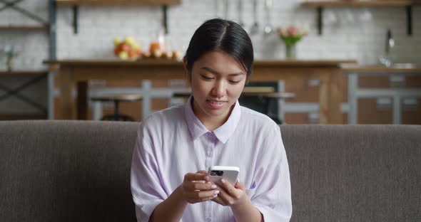 Attractive Asian Woman Sitting of the Sofa at Home Using Mobile Phone and Smiling