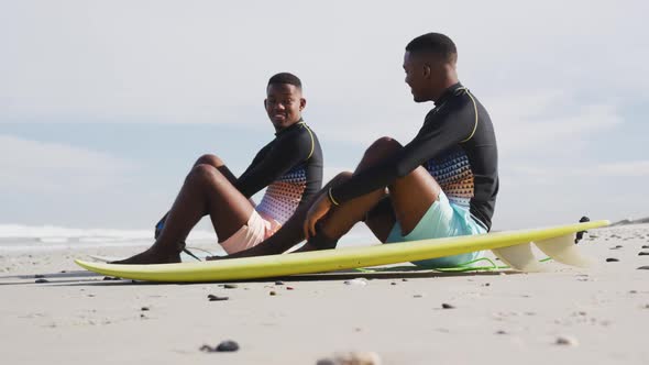 Happy african american teenage twin brothers sitting by surfboards on a beach talking