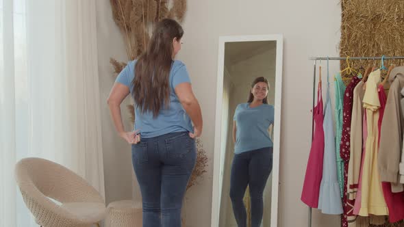 Cheerful Plump Woman in Jeans Admiring Reflection in Mirror Indoors