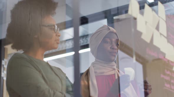 Two smiling african american businesswomen brainstorming using memo notes on glass wall in office