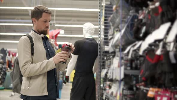 Sportive Man is Regarding a Plastic Bottle for Water in a Sport Store