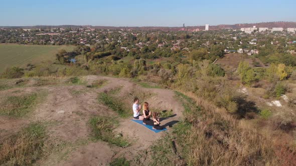 Orbiting Aerial Shot of Family of Three Mother Father and Daughter Do Yoga Exercises on Top of Hill