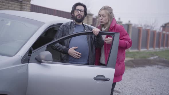 Caucasian Blond Woman Helping His Pregnant Husband To Get Into the Car