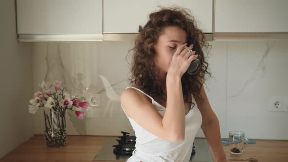 A Young Woman is Standing in the Kitchen and Drinking Coffee