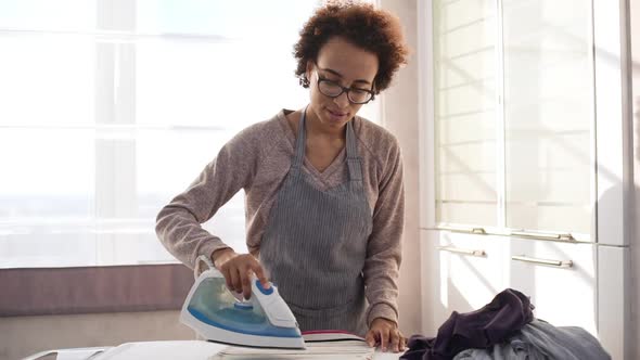 Young African American Female in Apron and Glasses Ironing Clothes on Board in Light Room