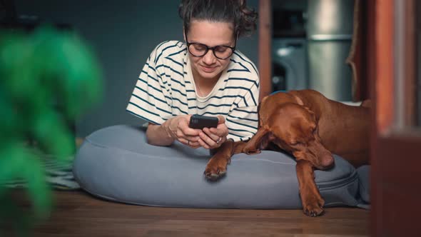 A Young Woman Using Her Smartphone While Lying Next to Her Cute Sleeping Dog