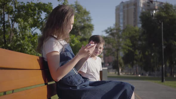 Older Sister with Younger Brother Sitting on the Bench in the Summer Park