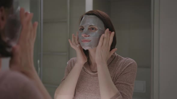Young Adult Woman Wearing Face Cosmetics Mask and Admiring with Her Reflection at Bathroom Mirror